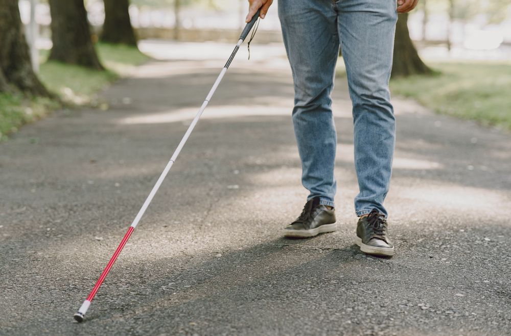 Imagem mostra uma pessoa com deficiência visual caminhando com uma bengala em rua