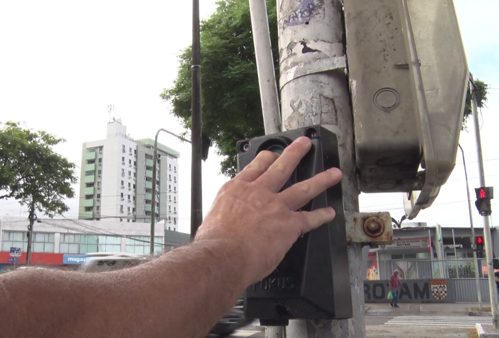 Na imagem, a mão de uma pessoa aparece acionando uma botoeira sonora de um semáforo